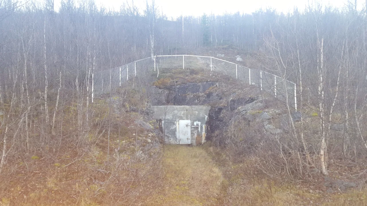 Inngjerdet fjellhule med en hvit dør og nettinggjerde langs toppen, omgitt av spredte trær.