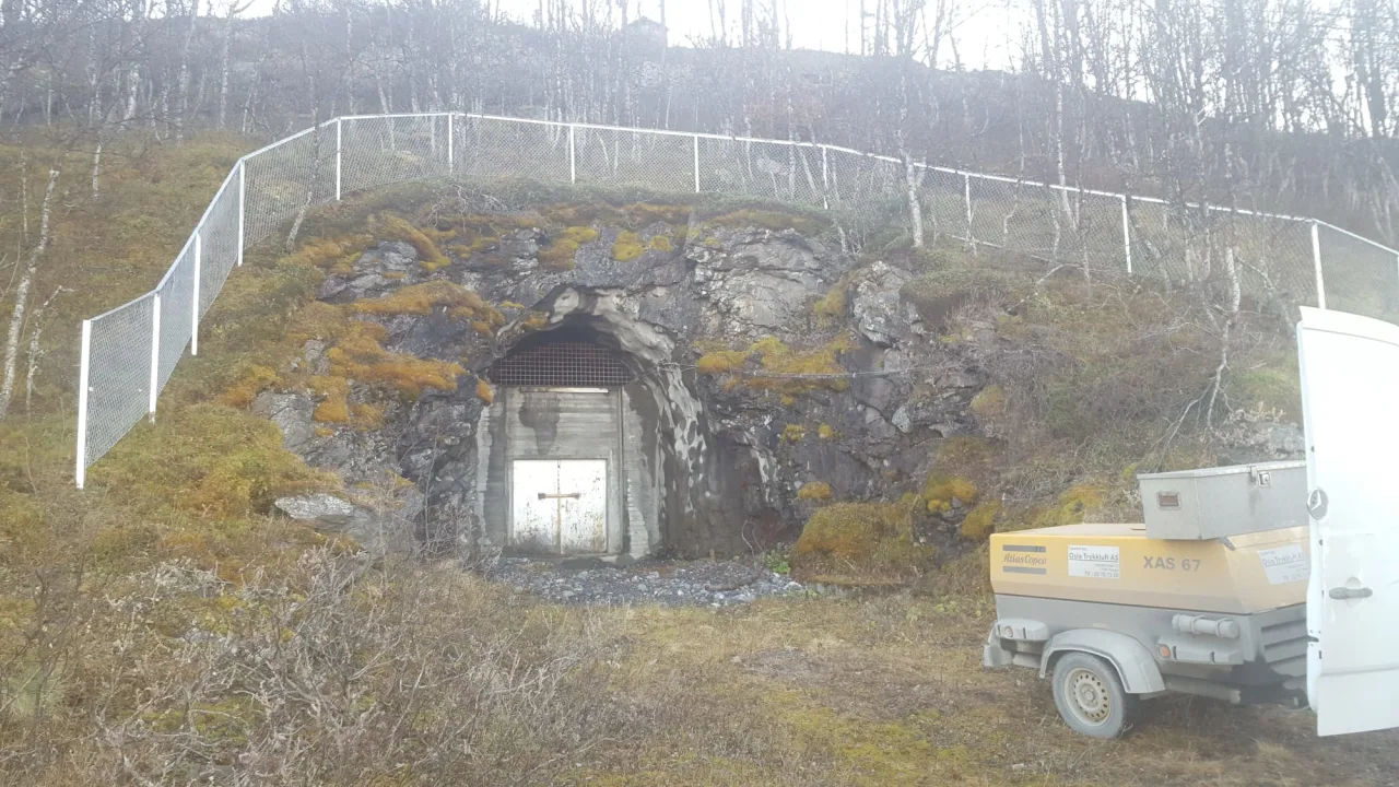 Fjellåpning med en port foran en betongdør, omgitt av mose og nettinggjerde på toppen av skråningen.