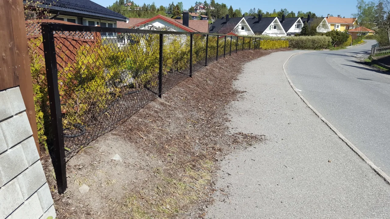 Nettinggjerde langs en asfaltvei, med hekk og bolighus i bakgrunnen på en solrik dag.