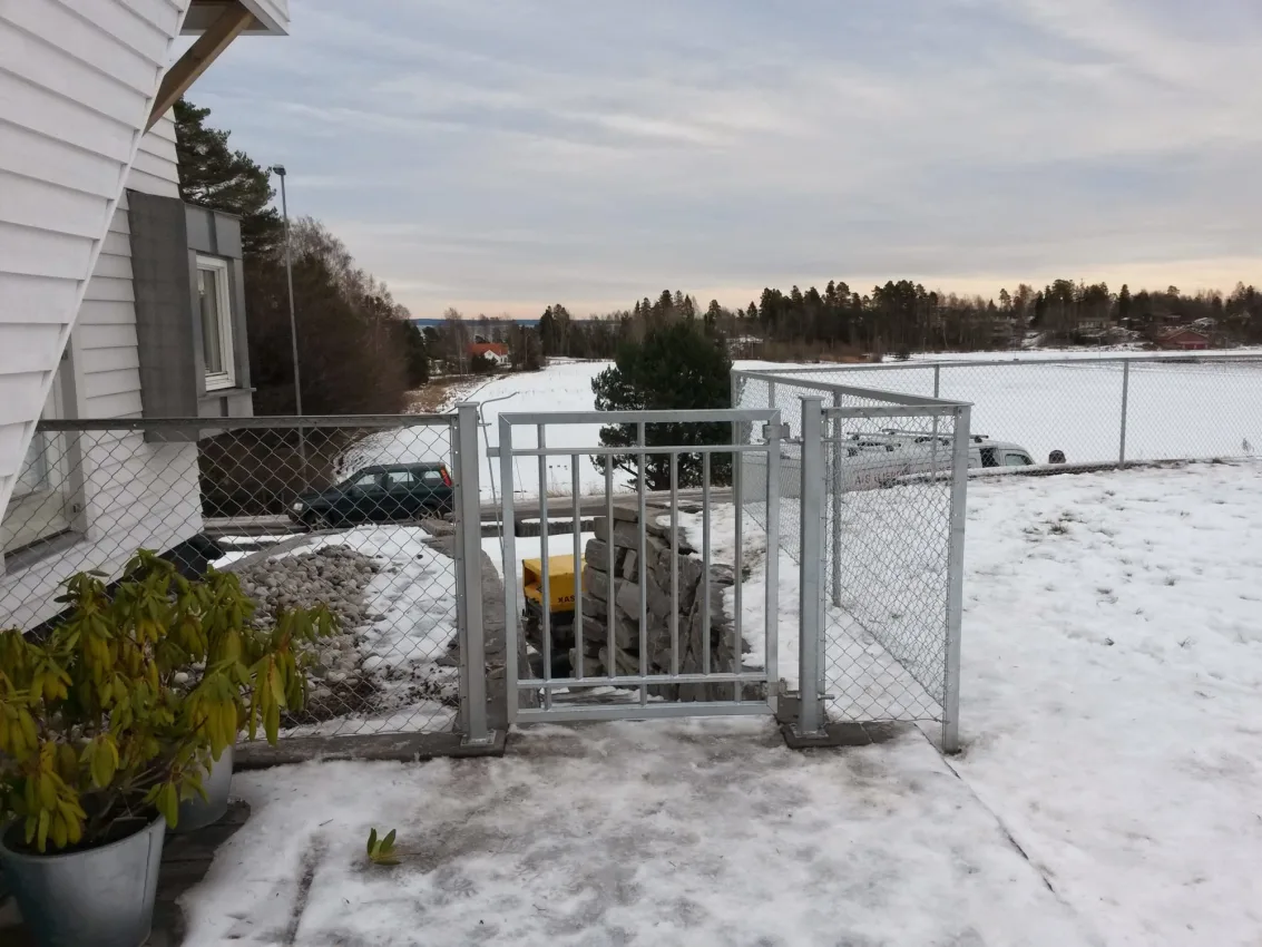 Metallport med nettinggjerde i vinterlig hage, med utsikt mot et åpent landskap og parkerte biler.