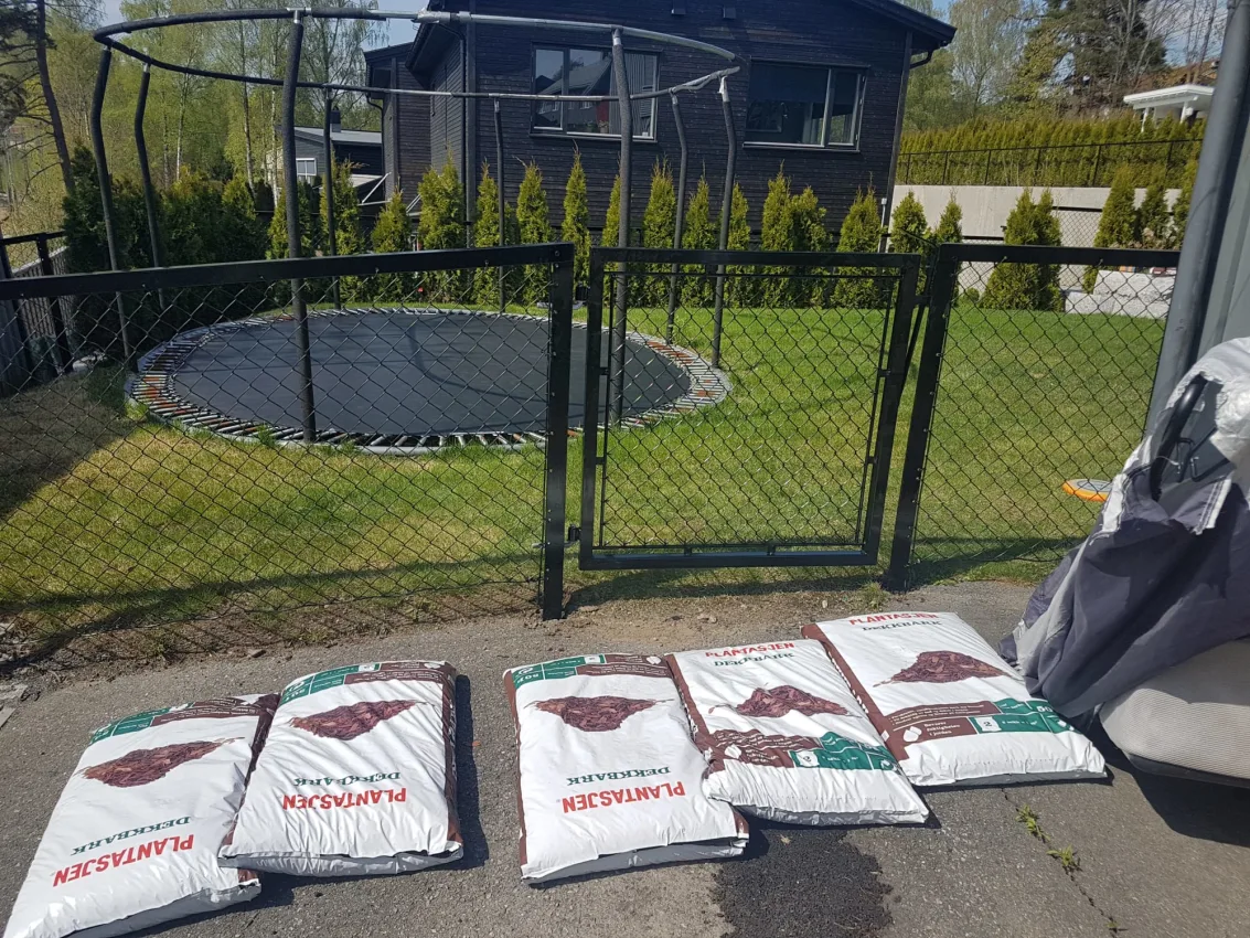 Metallport med nettinggjerde foran en gressplen og trampoline, med sekker hagebark i forgrunnen.